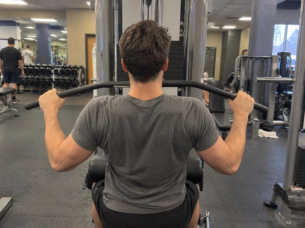 View from behind of a person seated at a lat pulldown machine in an ordinary gym, gripping the wide bar overhead.