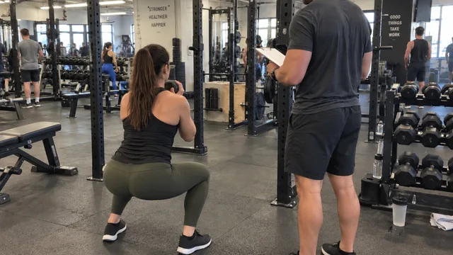 A personal trainer with a clipboard watching a client do a dumbbell goblet squat in an ordinary gym, both seen from behind.
