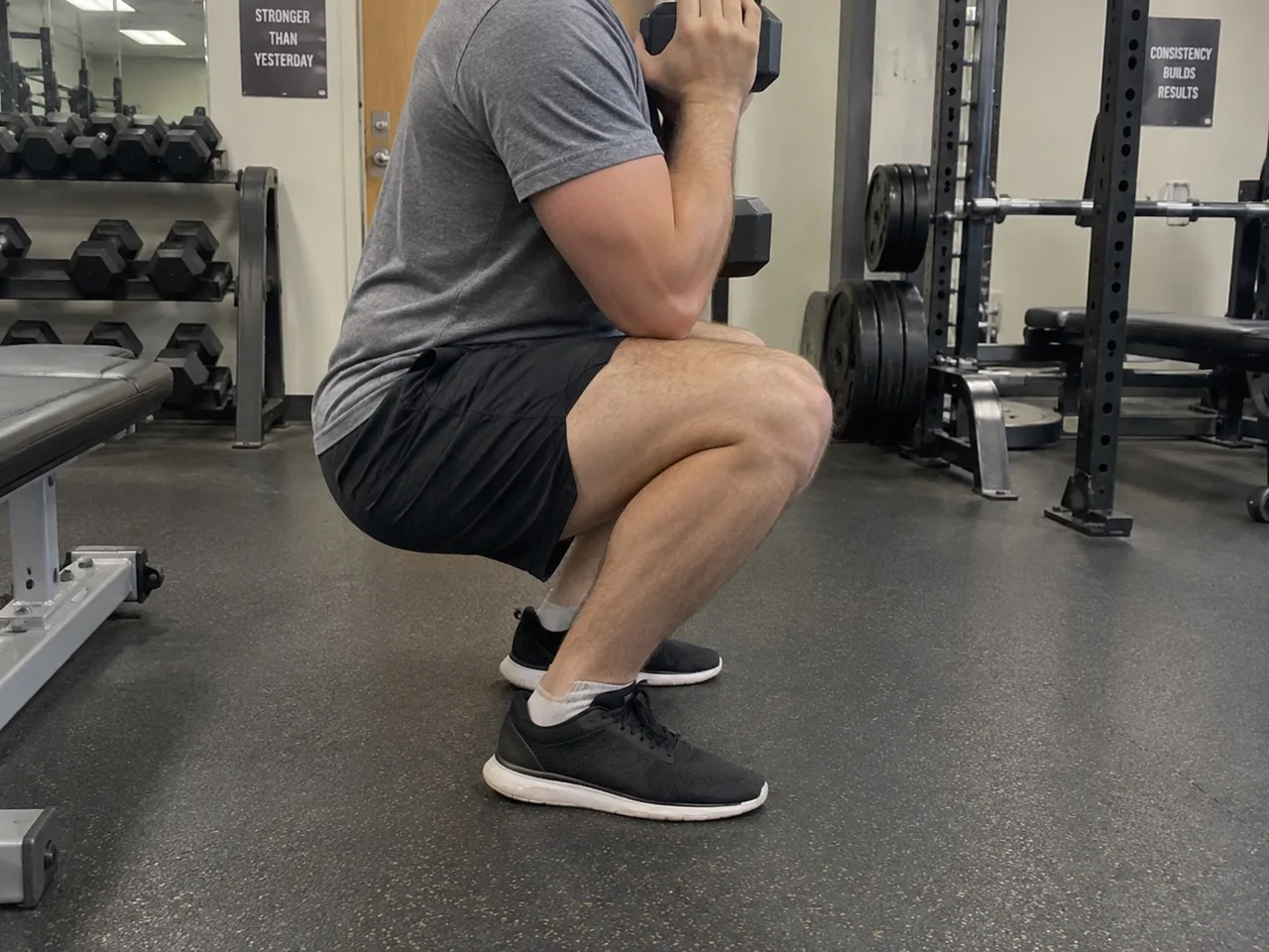 Side view of a person at the bottom of a goblet squat in a gym, holding a dumbbell vertically against their chest with heels flat on the floor.