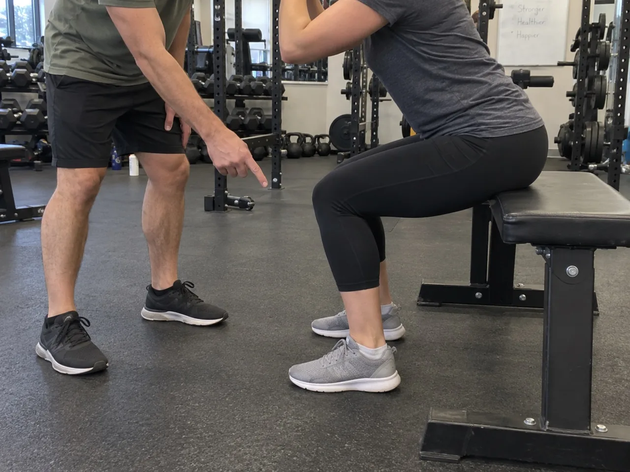A trainer standing beside a client doing a bodyweight box squat to a bench, pointing at the client's knees while giving a technique cue.