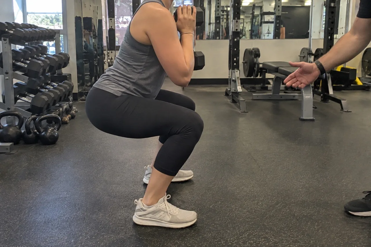 A beginner holding a single dumbbell at chest height during a light goblet squat in a gym, seen from the side with a trainer's hand gesturing at the edge of the frame.