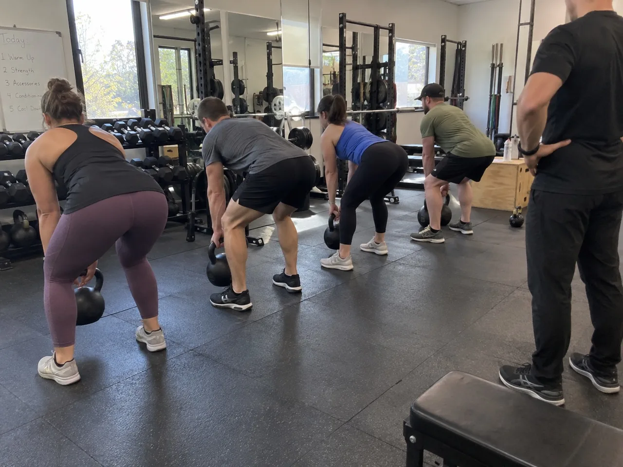 A small group of people seen from behind doing kettlebell deadlifts in a gym while a coach watches from the side, faces not visible.