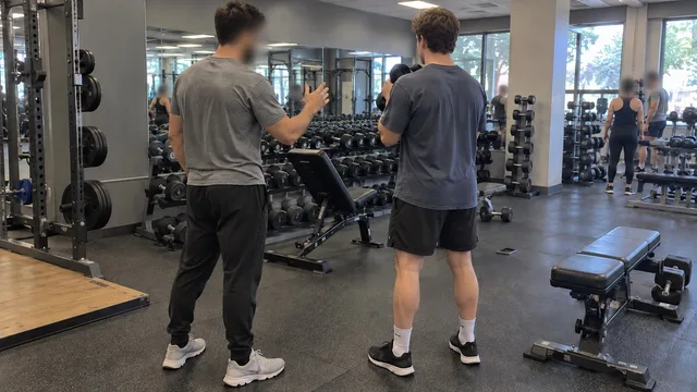 A personal trainer gesturing as he explains a movement to a client holding a dumbbell in a gym, both seen from behind.