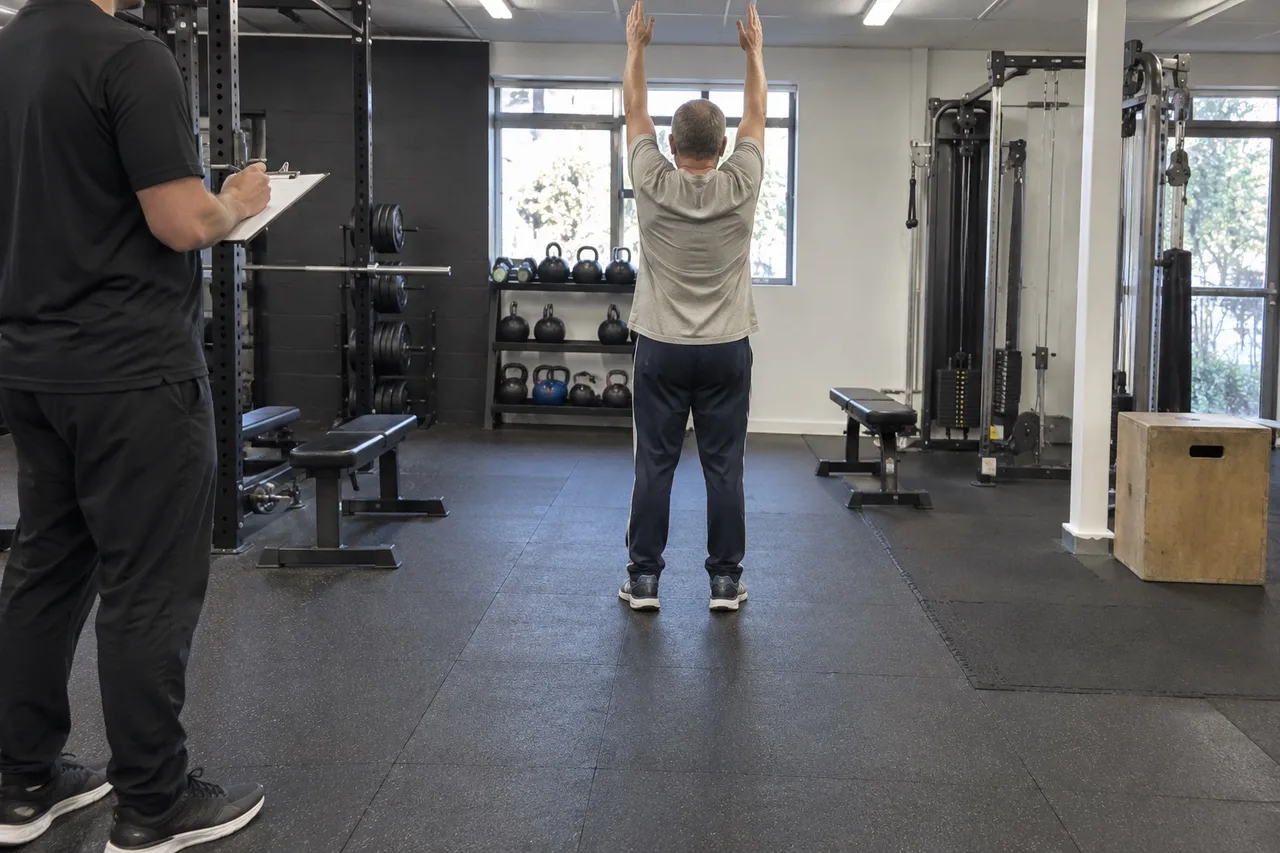 A beginner in a grey t-shirt and track pants reaching both arms overhead in a gym, seen from behind, while a trainer holding a clipboard watches from the side.