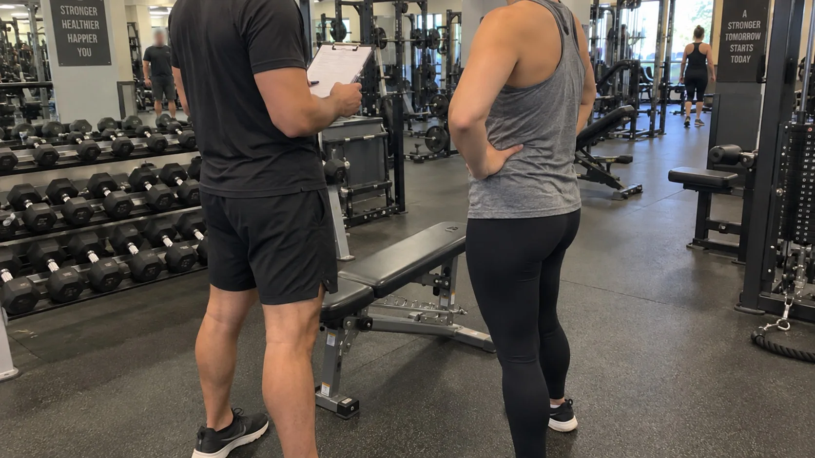 A personal trainer holding a clipboard talks with a new client next to a bench in a busy gym.