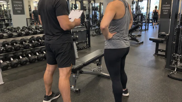 A personal trainer holding a clipboard talks with a new client next to a bench in a busy gym.