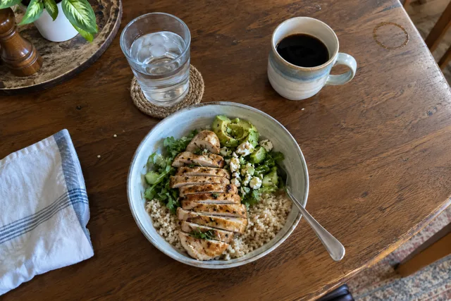 Overhead view of a grilled chicken, rice and greens bowl with a glass of water and a coffee on a wooden table.