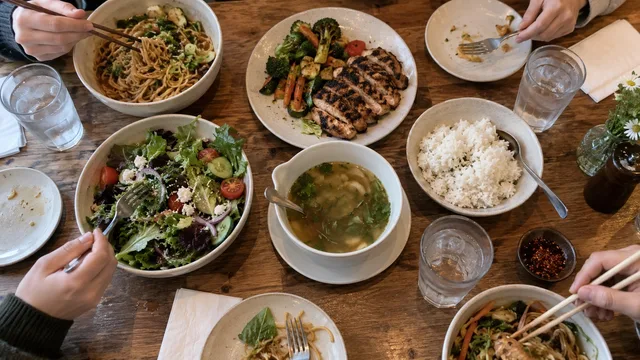 Overhead view of a shared table with noodles, rice, grilled chicken with vegetables, salad and soup, with hands reaching in with chopsticks and forks.