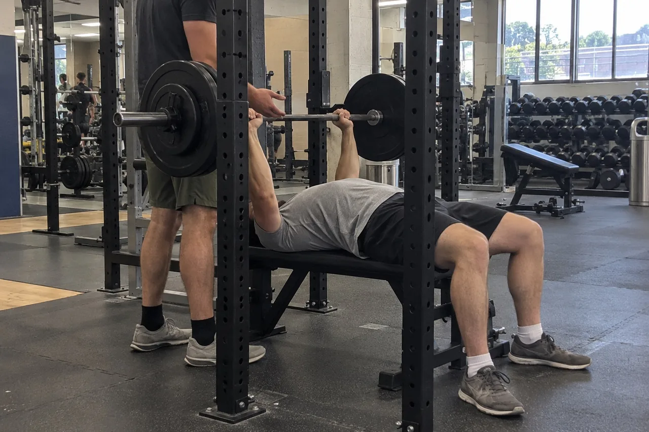 A person doing a barbell bench press while a trainer stands behind the bench with hands ready to spot the bar.