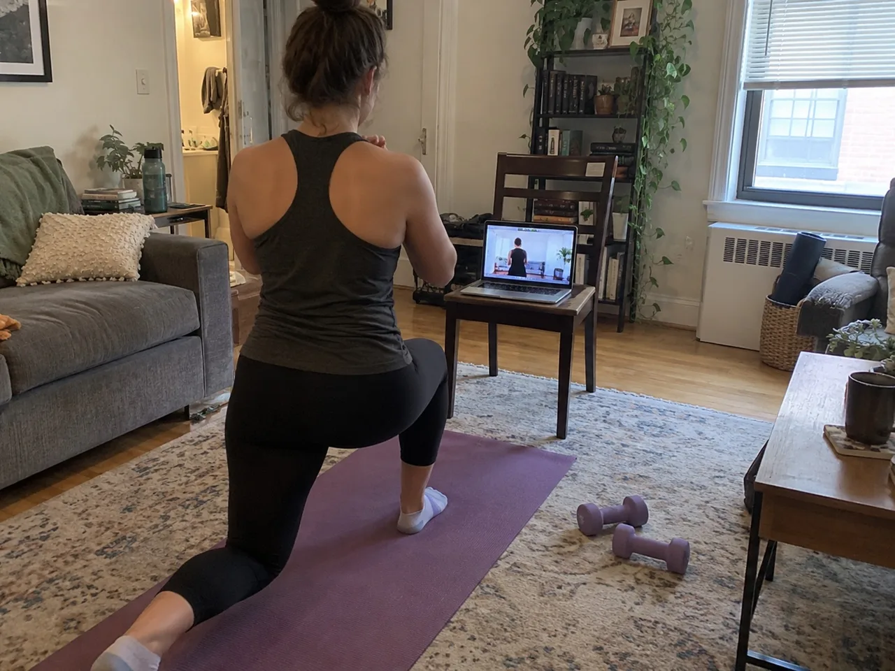 A person seen from behind doing a lunge on a yoga mat in a living room in front of a laptop on a chair showing a video call.