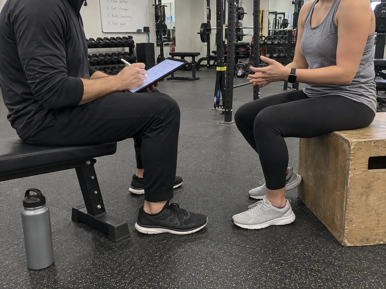 A trainer sitting on a gym bench taking notes on a tablet while talking with a new client during an initial assessment.