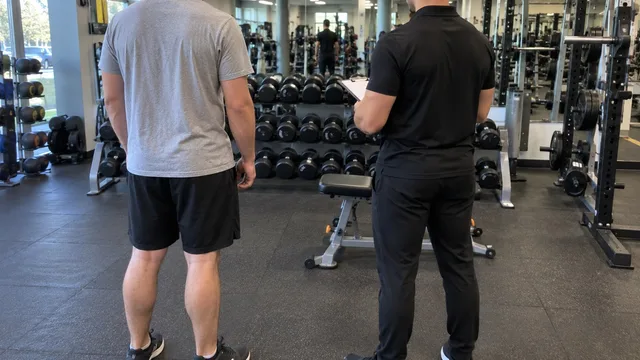 A new client in a grey t-shirt standing beside a personal trainer holding a clipboard, both seen from behind facing the dumbbell rack in a gym.