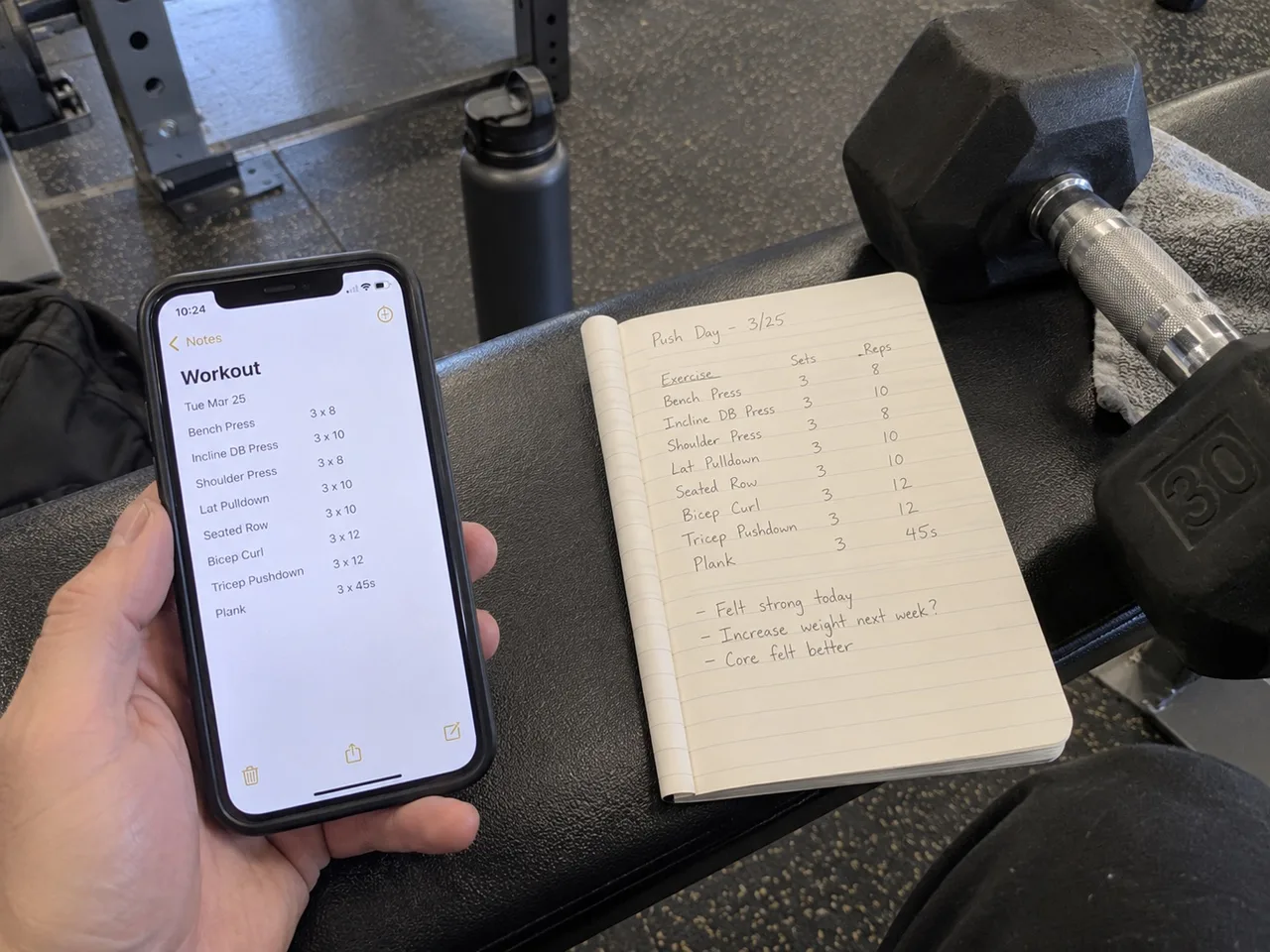 A hand holding a phone showing a workout log, next to an open notebook with handwritten exercises, sets and reps on a gym bench.